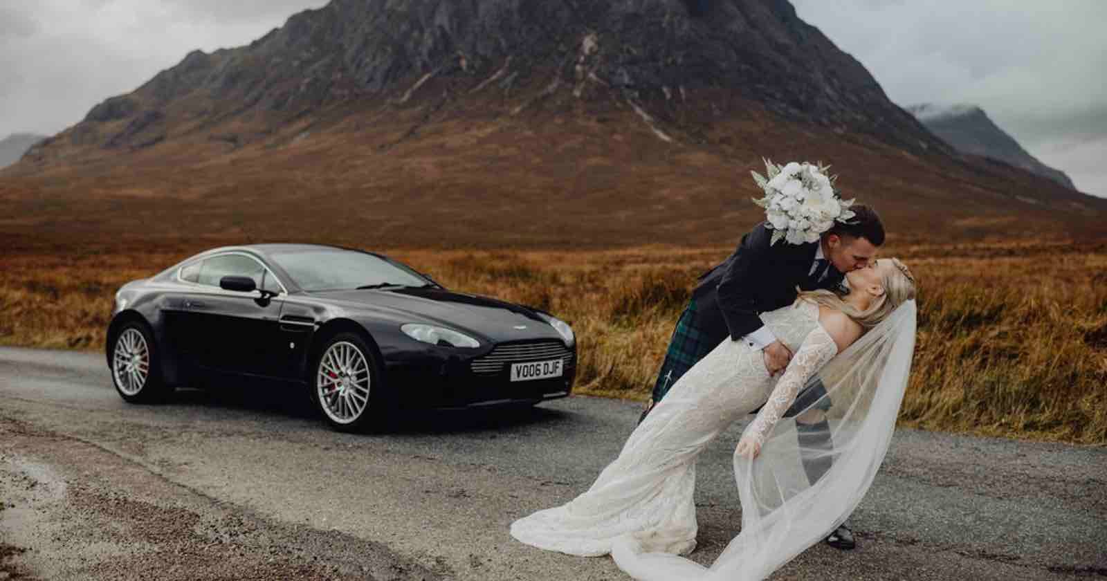 A groom dips and kisses his bride, who holds a bouquet and wears a long veil, on a road in front of a black sports car and a large mountain in a scenic, grassy landscape.