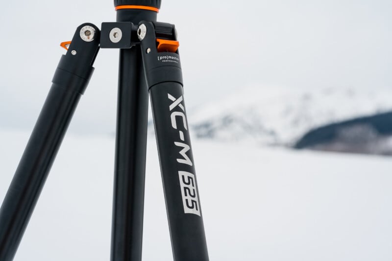 A close-up of a black tripod leg labeled “XC-M 525” standing on snow, with a blurred snow-covered mountain and cloudy sky in the background.