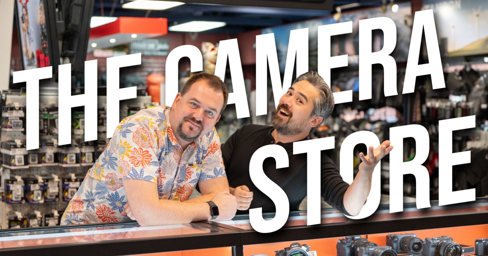 Two men stand behind a counter in a camera store, smiling at the camera. Large white text across the image reads "THE CAMERA STORE." Cameras and accessories are visible in the background.