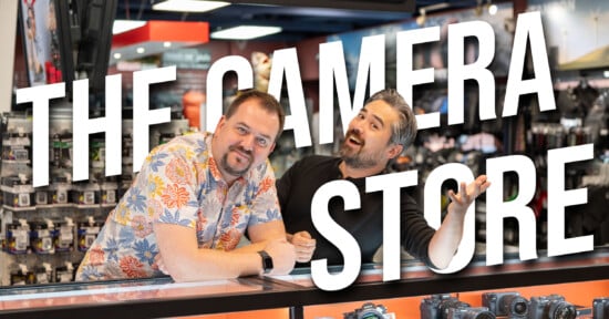 Two men stand behind a counter in a camera store, smiling at the camera. Large white text across the image reads "THE CAMERA STORE." Cameras and accessories are visible in the background.
