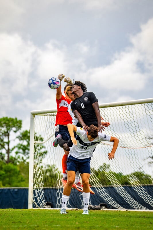 A soccer goalkeeper in an orange jersey jumps to punch the ball as two opposing players in black and white jerseys contest for a header in front of the goalpost. All three are mid-air and focused on the ball.