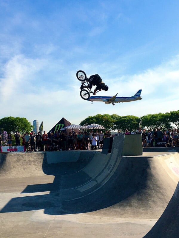 A BMX rider performs a high aerial trick at a skate park, with a crowd watching and an airplane flying low in the background against a clear blue sky.