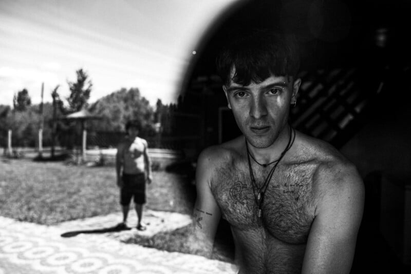 A shirtless young man with tattoos looks into the camera, partially in shadow. In the blurred background, another shirtless person stands outside on a sunlit, grassy area. The image is black and white.