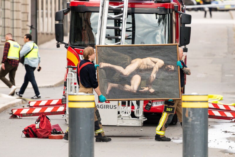 Two firefighters carry a large painting of a nude male figure in front of a fire truck on a city street, while other people in safety vests walk nearby.