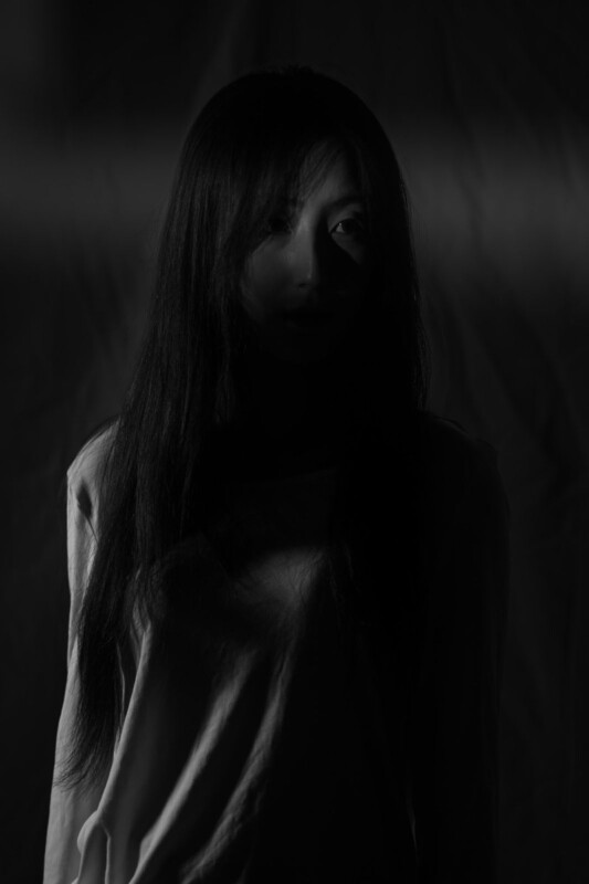 A person with long hair and a light-colored shirt stands in low, dramatic lighting, partially cloaked in shadow, with only part of their face and upper body faintly visible against a dark background.