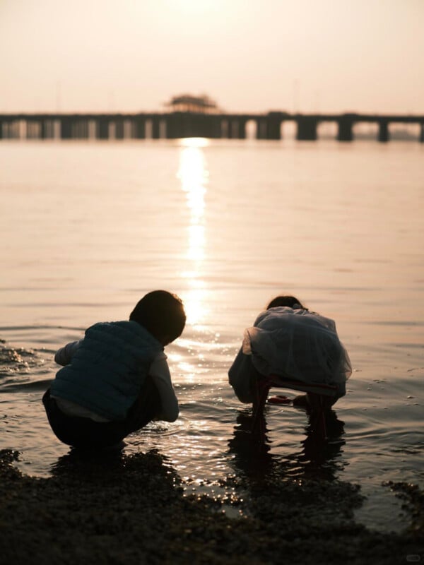 Two children crouch by the water's edge at sunset, silhouetted against the shining reflection of the sun on the calm surface, with a bridge visible in the background.