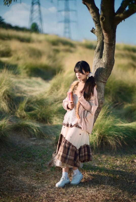A young woman in a long, layered dress and sweater leans against a tree, holding a book. She stands on grassy terrain with sunlight casting soft shadows, and power lines are visible in the background.
