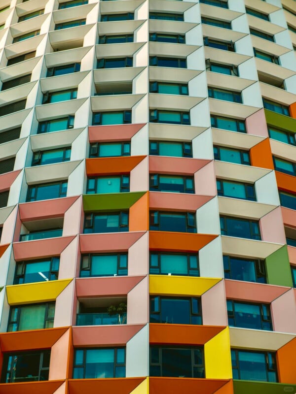 Colorful modern apartment building facade with geometric, three-dimensional window frames in shades of orange, yellow, pink, and white, arranged in a repeating pattern across multiple floors.