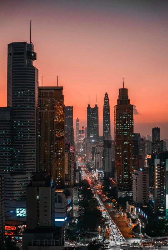 A city skyline at sunset with tall skyscrapers on both sides and a busy street filled with car headlights stretching into the distance under an orange-pink sky.