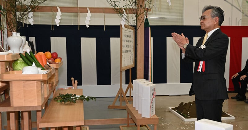 A man in a suit claps his hands in prayer at a traditional Japanese ceremony, facing an altar with offerings of fruits, vegetables, and sake bottles. Ritual objects and a mound of dirt are present in the background.