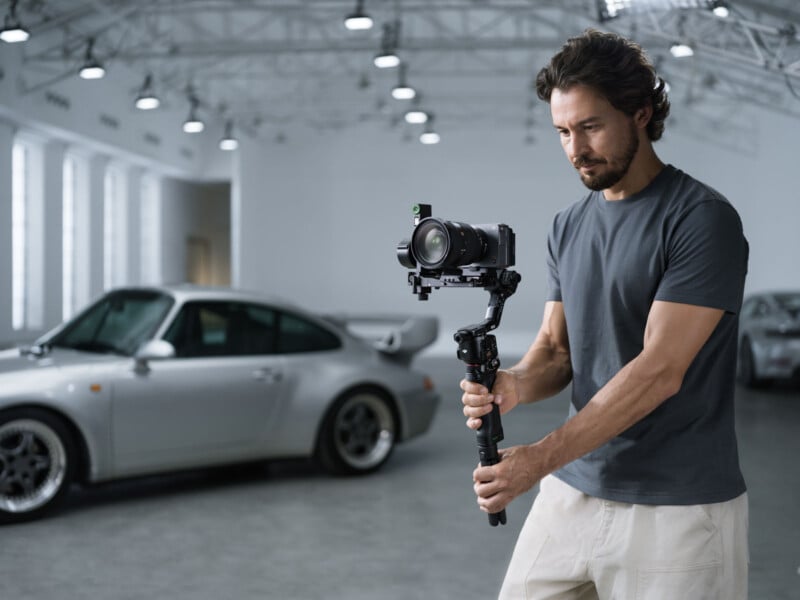 A man in a gray t-shirt operates a camera on a gimbal inside a spacious, well-lit garage with a silver sports car parked in the background.