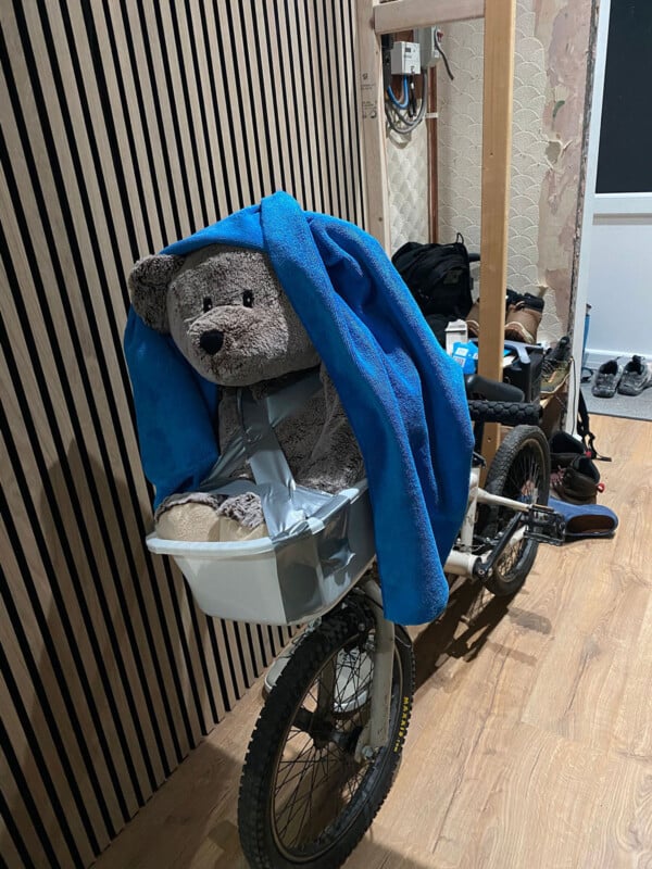 A large teddy bear covered with a blue towel sits in a plastic tub taped to the front of a bicycle indoors, near a wall with wooden slats and several pairs of shoes on the floor in the background.