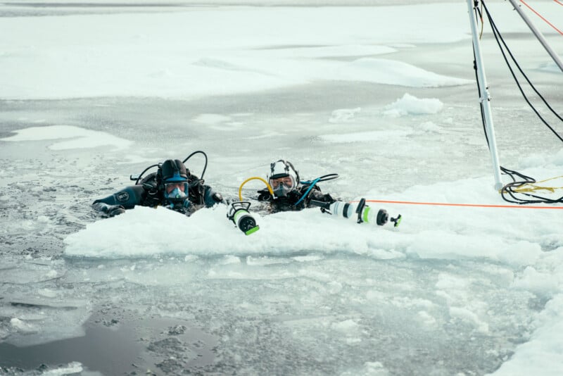 Two scuba divers in cold-weather suits emerge from a hole cut in the ice, surrounded by snow and icy water, with ropes and equipment visible nearby.