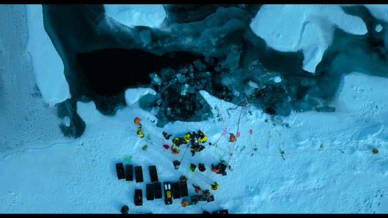 Aerial view of people in winter gear gathered around a hole in the ice near open water, with equipment and sleds arranged nearby on a snow-covered landscape.