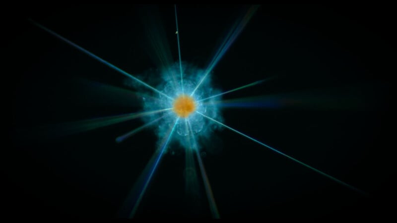 A glowing blue and orange particle emits bright, colorful rays against a black background, resembling an artistic depiction of an atom or a burst of energy in space.