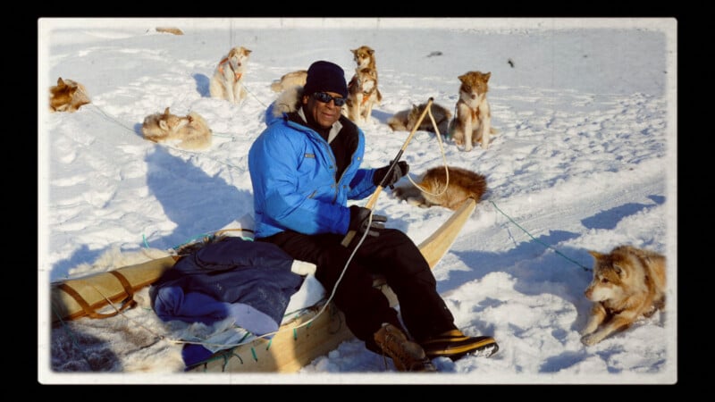 A person in a blue jacket and sunglasses sits on a wooden sled in the snow, holding a rope, surrounded by several sled dogs resting and sitting on the snowy ground.