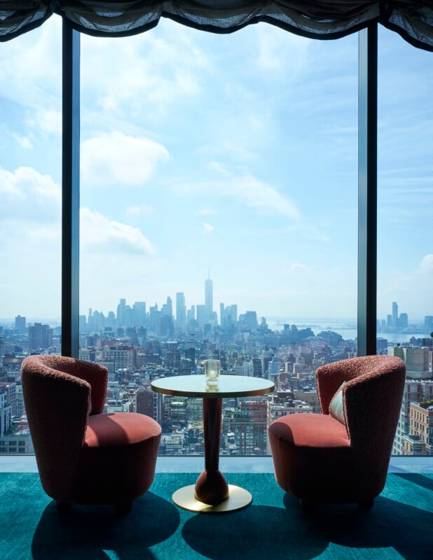 Dos sillones rosas y una pequeña mesa redonda con cristal se encuentran junto a un gran ventanal con vistas a un paisaje urbano de rascacielos y un cielo parcialmente nublado.