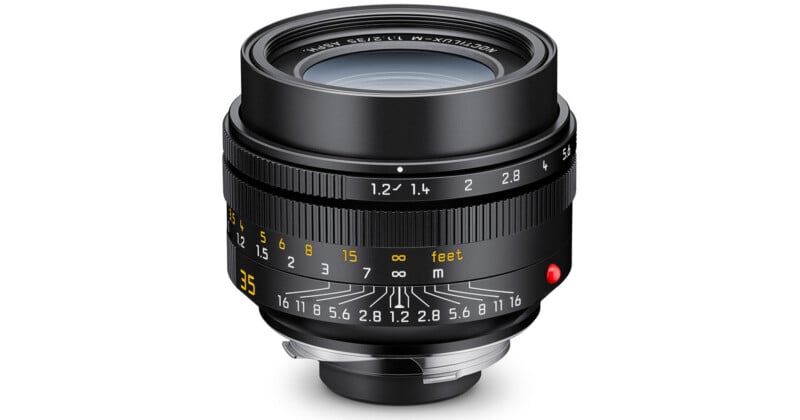 A close-up view of a black camera lens with detailed focus and aperture markings in white and yellow, a red indicator dot, and a silver mounting ring at the base.