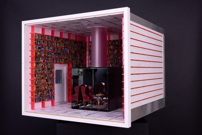 Maqueta de habitación en forma de cubo con paredes blancas, decoración roja y estantes coloridos. En el interior hay sillas rojas, mesas negras y una columna central rosa, creando un interior moderno y futurista sobre un fondo negro.