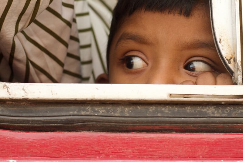 A young boy rests his chin on his hand and looks out of a window with a thoughtful expression. He is partially visible behind the window frame, and someone wearing a striped shirt is next to him.
