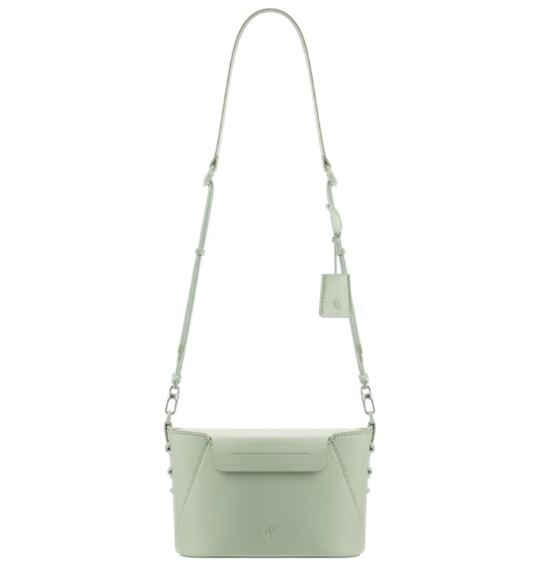 A minimalist light green leather crossbody bag with a rectangular shape, adjustable shoulder strap, small flap closure, and subtle stitched details. A small tag hangs from the strap.
