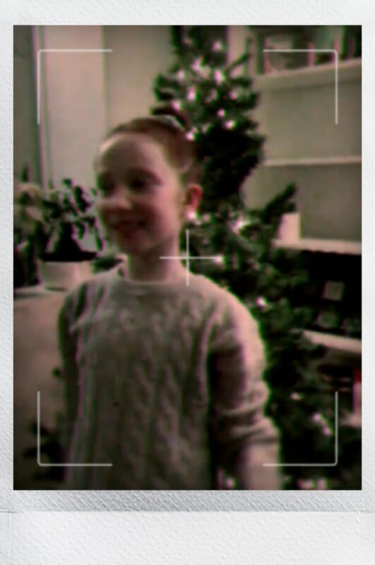 A young girl with red hair in a bun, wearing a light-colored sweater, stands smiling in front of a decorated Christmas tree indoors. The image appears slightly blurry and has visible camera focus lines.