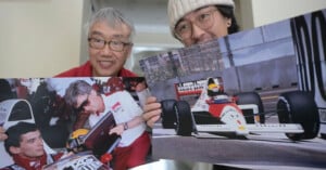 Two men smile at the camera while holding large photos of Formula 1 scenes, including a driver signing an autograph and a racing car on the track.