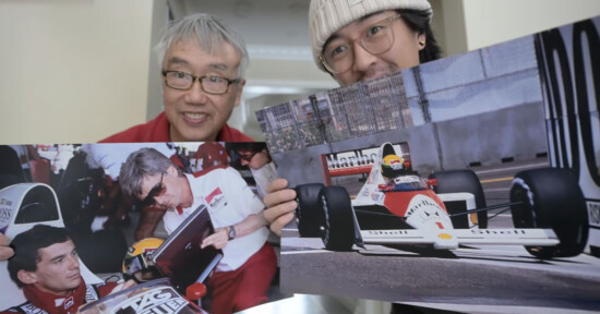 Two men smile at the camera while holding large photos of Formula 1 scenes, including a driver signing an autograph and a racing car on the track.