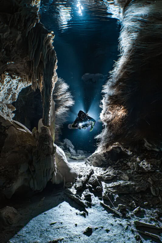 Un buzo, iluminado por un rayo de luz, explora una cueva submarina rodeada de espectaculares formaciones rocosas y estalactitas mientras burbujas se elevan hacia la superficie.