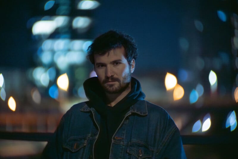 A man wearing a denim jacket and hoodie stands outdoors at night, with colorful blurred city lights in the background. The lighting is dim, giving the scene a moody atmosphere.