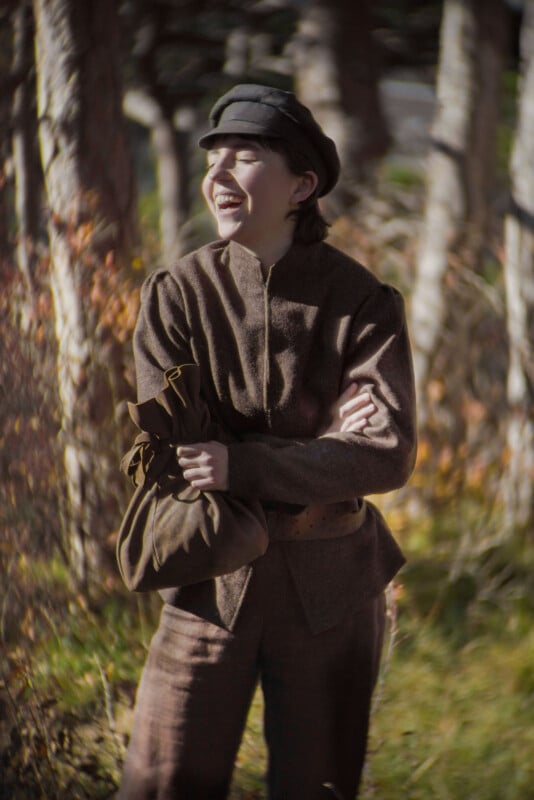 A person dressed in old-fashioned brown clothes and a black cap stands outdoors in a sunlit forest, smiling and laughing while holding a bundled bag under one arm.