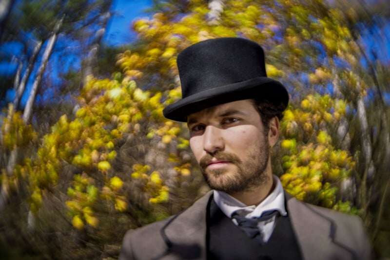 A man with a beard wearing a black top hat and vintage suit stands outdoors with blurred yellow foliage and blue sky in the background, creating a swirling focus effect.
