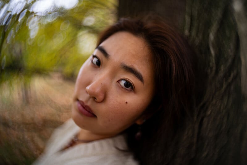 A woman with medium-length brown hair leans against a tree, looking softly at the camera. The background is blurred with swirling green and brown tones, creating a dreamy effect.