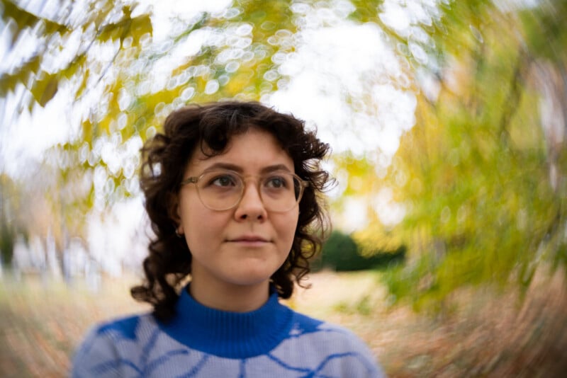 A person with curly hair and glasses stands outdoors, surrounded by blurred autumn foliage and a bright, swirling bokeh background. They wear a blue and white sweater and look slightly off to the side, appearing thoughtful.