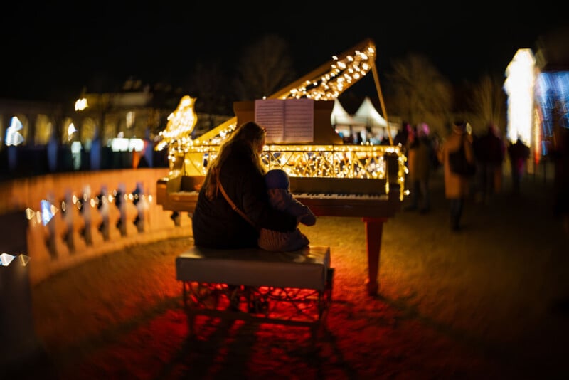 A person and a child sit together at a piano decorated with string lights, outdoors at night, with blurred people and glowing lights in the background.