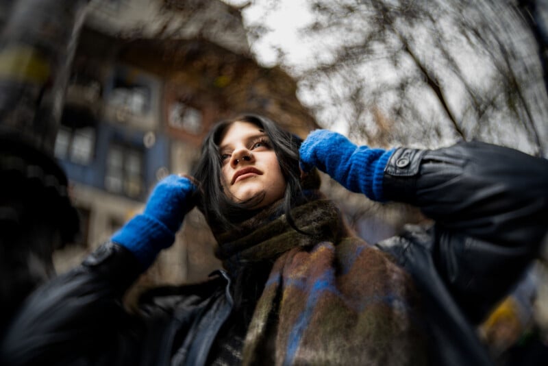 A woman wearing blue gloves, a black coat, and a scarf stands outdoors, looking up. The background is blurred with buildings and bare trees, creating a moody, wintry atmosphere.