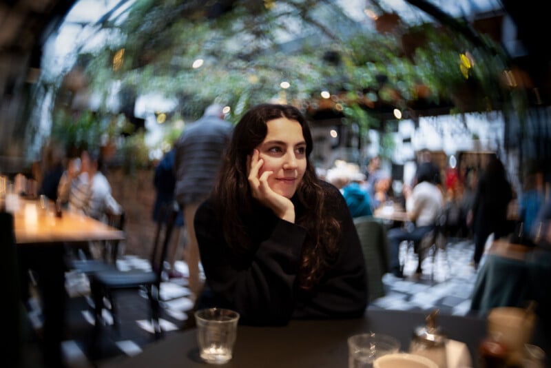 A woman with long dark hair sits at a table in a lively, blurred cafe with greenery overhead, resting her chin on her hand and smiling softly, with empty glasses and cups in front of her.