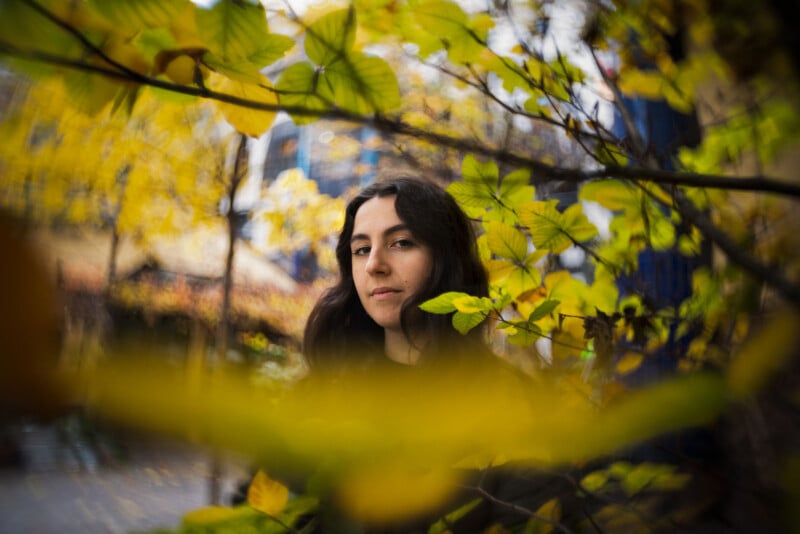 A woman with long dark hair looks at the camera through yellow autumn leaves, with blurred branches in the foreground and a cityscape in the background.