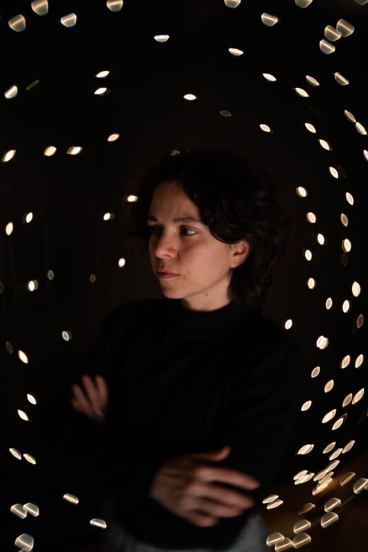 A person with short dark hair and a black top stands with arms crossed, surrounded by swirling, out-of-focus lights on a dark background, creating a dreamy, contemplative atmosphere.