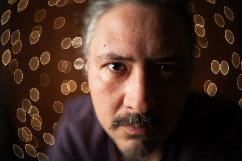 A close-up of a man with a mustache and beard, looking intently at the camera. The background is filled with warm, circular bokeh lights, creating a soft, artistic effect.