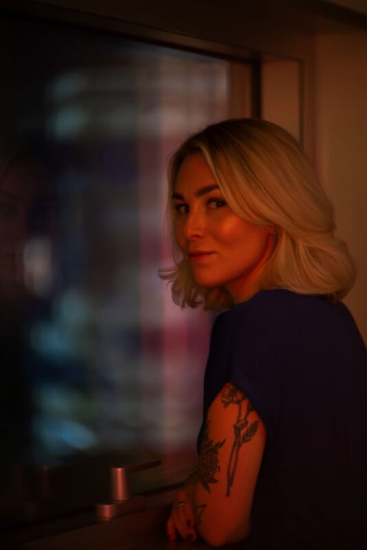A woman with blonde hair and a floral tattoo on her arm gazes softly over her shoulder, standing by a window with dim, warm lighting and blurred reflections in the glass.