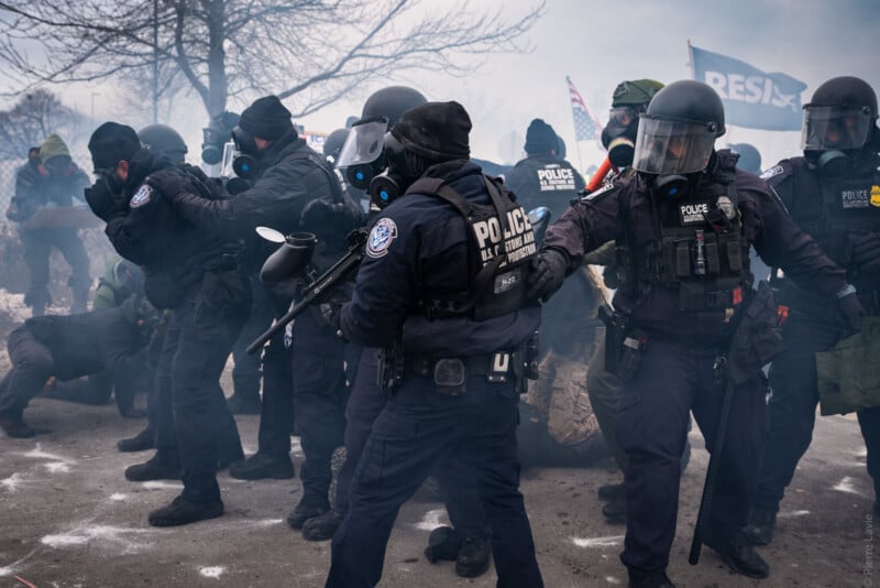 Police officers in riot gear and gas masks confront a crowd in a smoky, tense outdoor scene with American flags and a "RESIST" sign visible in the background. Some people are crouching or on the ground.