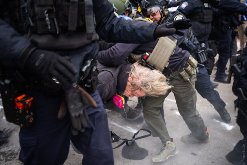 A person with light hair is being detained by police officers dressed in dark uniforms and tactical gear during a chaotic scene on a street. The person is bent over, and their mask is hanging from their neck.