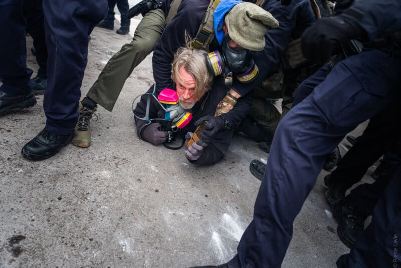 A man wearing a gas mask and a colorful respirator is pinned to the ground by police officers, surrounded by their legs and boots on a concrete surface.