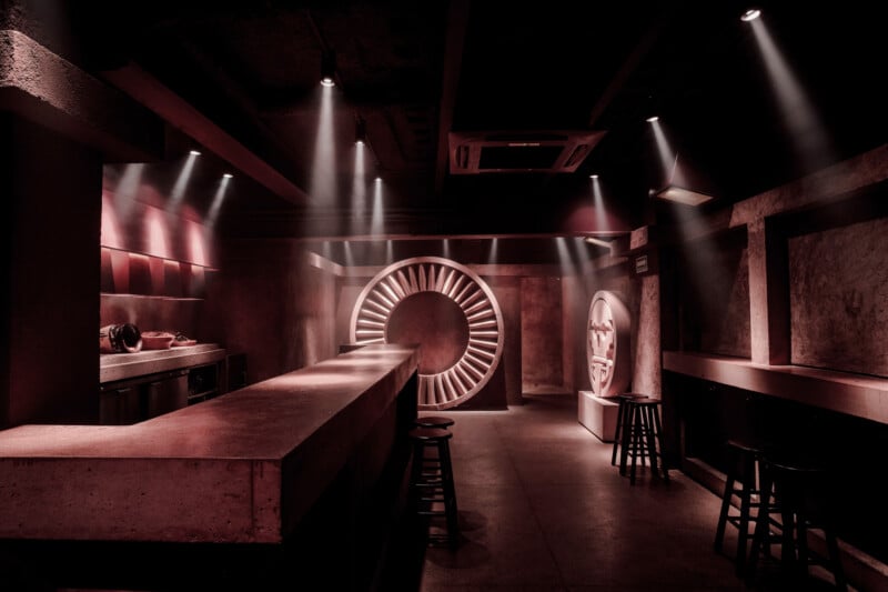 Barra con poca luz, luces rojas, mostrador de concreto, taburetes negros y una puerta redondeada en forma de bóveda en la parte de atrás. Los focos crean sombras dramáticas y le dan al espacio un ambiente industrial y misterioso.