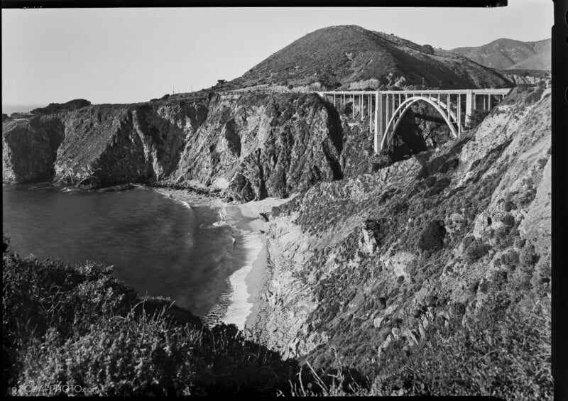 Black-and-white photo of a tall arched bridge spanning rugged coastal cliffs above the ocean, with waves crashing on a narrow beach below and hills rising in the background.