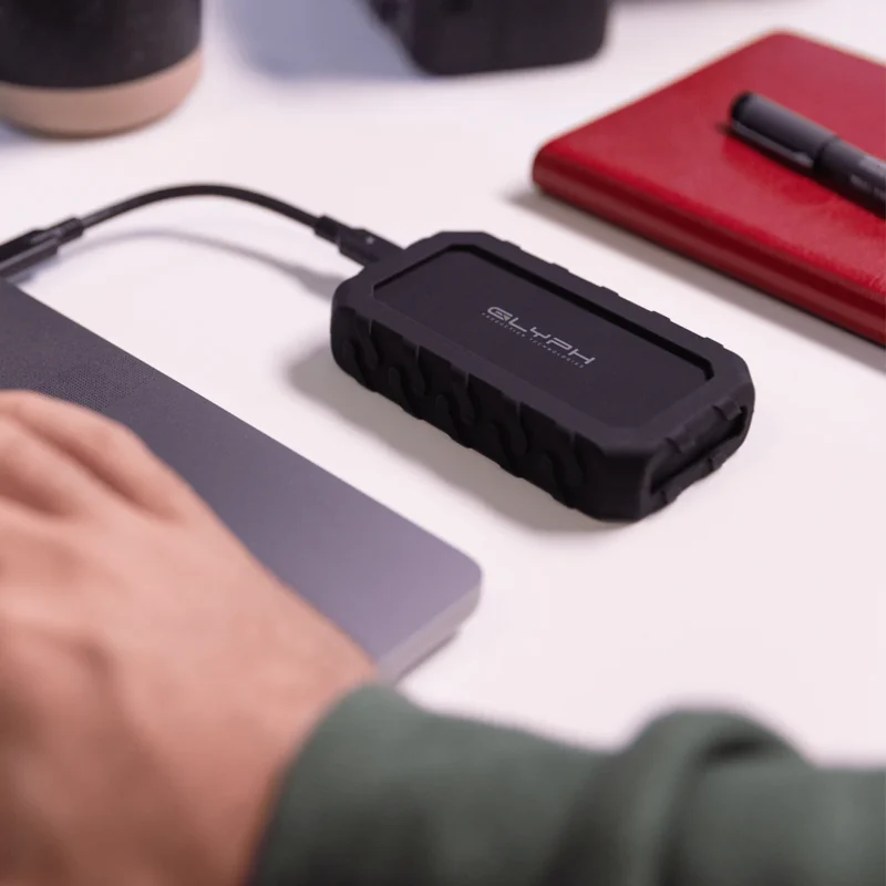 Las manos de un hombre usando una computadora portátil conectada a un disco duro externo Glyph sobre una mesa blanca, junto a una libreta roja y un bolígrafo negro.