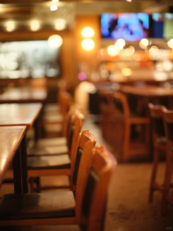 A row of empty wooden chairs and tables in a warmly lit restaurant or bar, with the background out of focus, showing blurred lights and a TV screen.