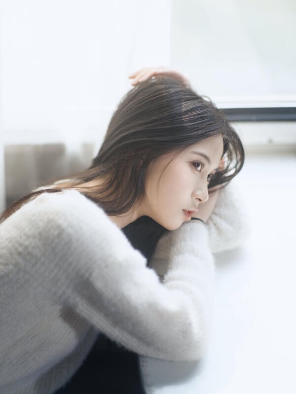 A young woman with long dark hair sits by a bright window, resting her head on her arm and gazing thoughtfully into the distance, wearing a soft, light-colored sweater.