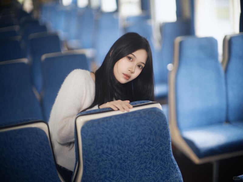 A young woman with long black hair and a white sweater sits alone in a row of blue patterned seats on a bus or train, looking back over the seat in front of her. The background is softly blurred.